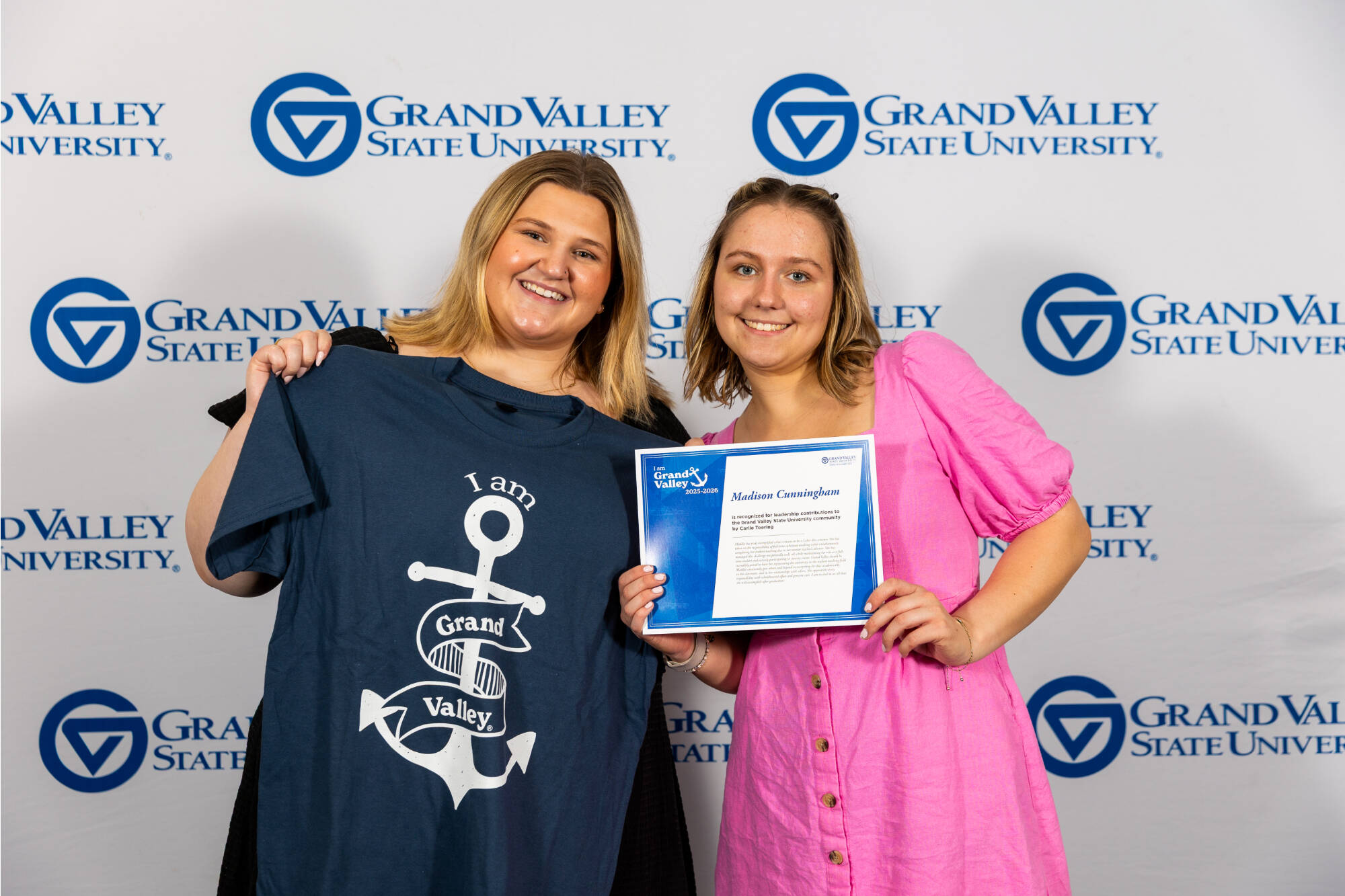 Two students smiling for the camera and holding up an "I am Grand Valley" t-shirt and an award certificate